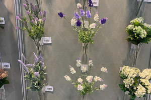 Artificial flowers displayed infront of a grey wall. The flowers are Lilac, Purple and white with green stems and leaves.