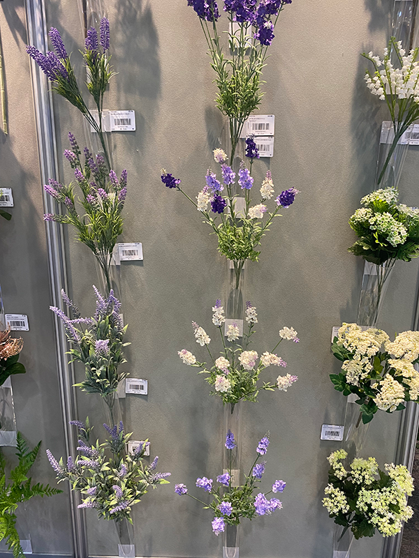 Artificial flowers displayed infront of a grey wall. The flowers are Lilac, Purple and white with green stems and leaves.